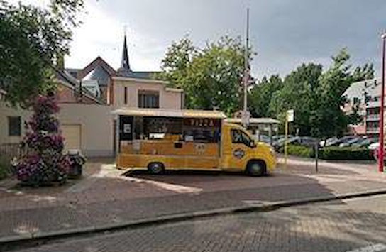 Geel voedseltruck geparkeerd op een plein met bomen en gebouwen op de achtergrond. (vergroot foto)