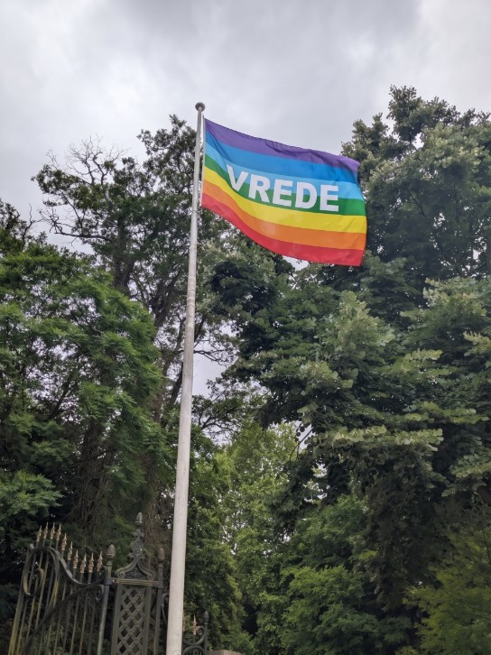 Regenboogvlag met het woord "VREDE" wappert aan een vlaggenmast tussen groene bomen. (vergroot foto)