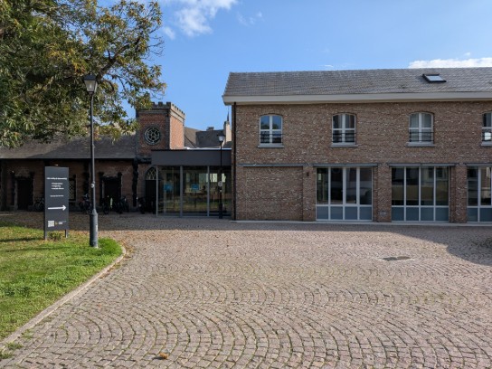 Oud bakstenen gebouw met grote ramen en een kloktoren, omgeven door een geplaveid plein en bomen. (vergroot foto)