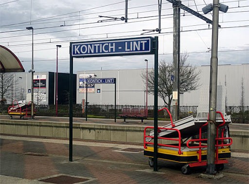Station Kontich-Lint met borden waarop de naam van het station staat, een leeg perron en een karretje met metalen platen en rode handgrepen onder een bewolkte hemel. (vergroot foto)